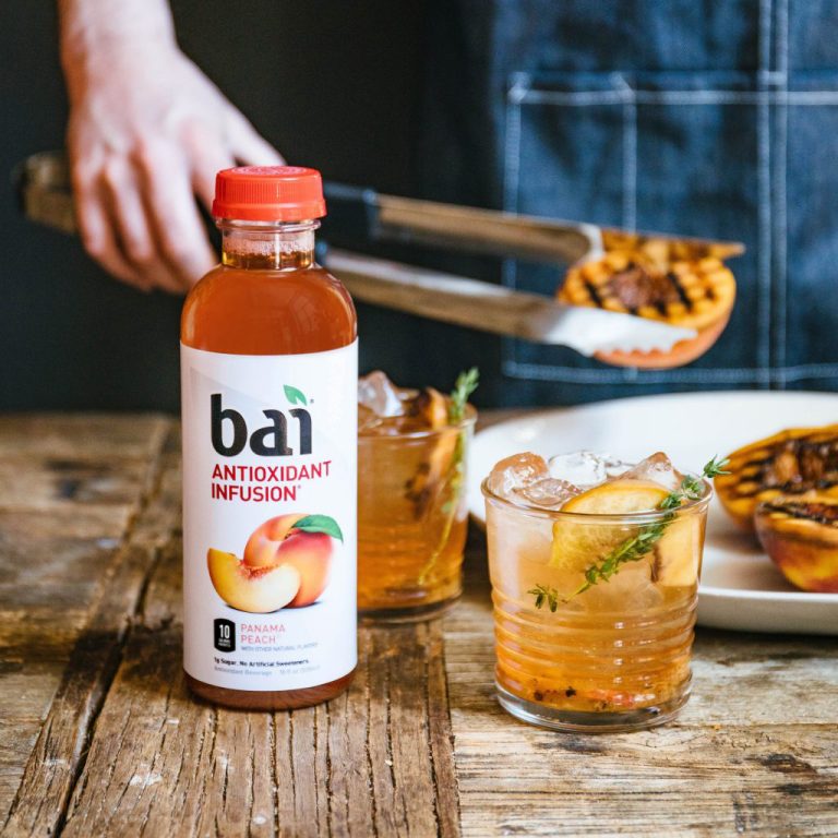 Man holding grilled peach with tongs by a Bai Panama Peach cocktail